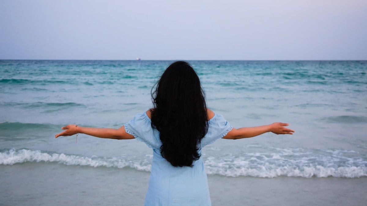 A woman stands at the edge of the ocean, arms outstretched, embracing the vast and calming waters in front of her.